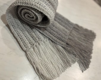 Könnte beinhalten: Ein gerollter, handgestrickter Schal in Grautönen mit Fransen. Der Schal ist aus weichem, strukturiertem Garn gefertigt, und die Farbe geht von Hell- zu Dunkelgrau über. Die Fransen sind in einem helleren Grauton gehalten.