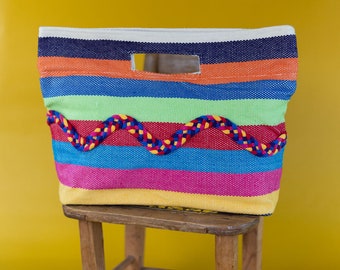 Puede incluir: Una bolsa de mano de rayas coloridas con un borde trenzado se encuentra sobre un taburete de madera. La bolsa tiene un asa y está hecha de tejido tejido.