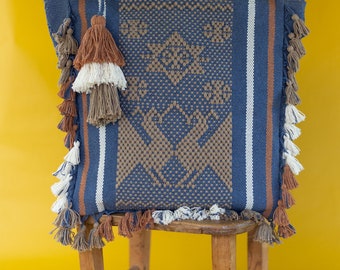 Puede incluir: Una bolsa de mano tejida azul y marrón con flecos. La bolsa está sentada en un taburete de madera sobre un fondo amarillo.