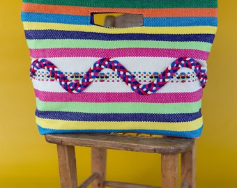 Puede incluir: Una bolsa de mano de rayas coloridas con un diseño tejido. La bolsa está sentada en un taburete de madera.