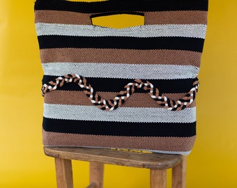 Puede incluir: Un bolso tote a rayas con un esquema de color marrón, blanco y negro. El bolso tiene un diseño tejido y un ribete trenzado marrón. El bolso está sentado en un taburete de madera.