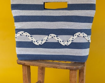 Puede incluir: Una bolsa de mano de rayas azules y blancas con un ribete ondulado blanco. La bolsa está sentada en un taburete de madera.