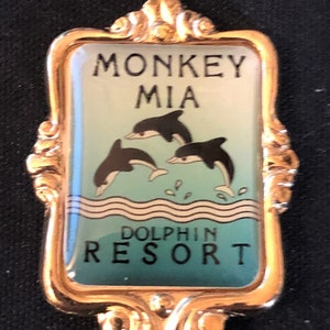 May include: A gold-colored souvenir spoon with a blue and white enamel design. The spoon features a graphic of three dolphins jumping out of the water. The text "Monkey Mia Dolphin Resort" is printed on the spoon.