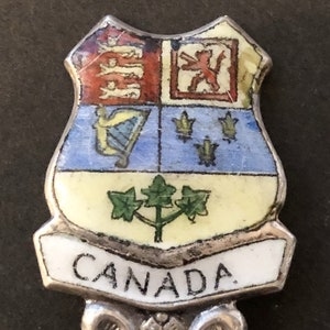 Op de afbeelding: Een zilveren souvenirlepel met een kleurrijk emailleontwerp van het Canadese wapen. De lepel heeft een decoratief handvat en het woord "CANADA" staat op de voorkant.