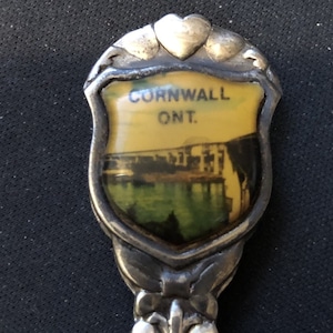 May include: Silver souvenir spoon with a shield-shaped charm featuring a picture of a town and the text "Cornwall Ont."