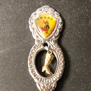 May include: Silver souvenir spoon with a gold-colored charm of a cowboy boot hanging from the handle. The handle is decorated with a shield-shaped emblem that says "TEXAS" and depicts a cowboy riding a horse.