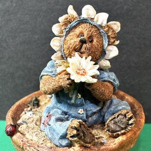 Può includere: Una statuetta di orso marrone con un abito in denim blu e un cappello a forma di girasole, che tiene un fiore bianco. L'orso è seduto in un vaso di fiori marrone con una coccinella sul lato. L'oggetto è decorativo.