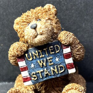 May include: A brown teddy bear figurine holding a sign that reads "UNITED WE STAND." The sign is blue with white stars and red and white stripes. The bear has detailed fur and paw prints.
