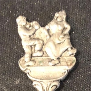 Puede incluir: Cuchara de plata con un mango adornado y un cuenco. El mango presenta un diseño esculpido de dos figuras.