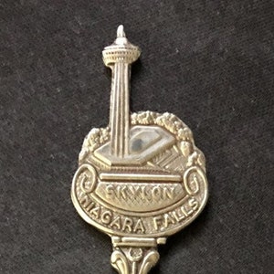 Op de afbeelding: Zilveren souvenirlepel met een gedetailleerd ontwerp van de Skylon Tower in Niagara Falls, Ontario, Canada. De steel heeft een decoratief ontwerp en de kom is rond en glad.