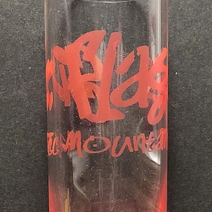 May include: A clear glass shot glass with a red inscription that reads "Surf's Up" and "Team Mountain".