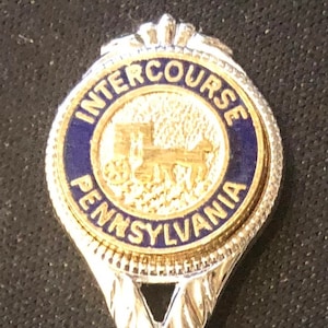 Könnte beinhalten: Ein silberner Löffel mit einem runden blauen und goldenen Anhänger. Der Anhänger trägt die Worte "INTERCOURSE PENNSYLVANIA" und ein goldenes Bild einer Pferdekutsche.