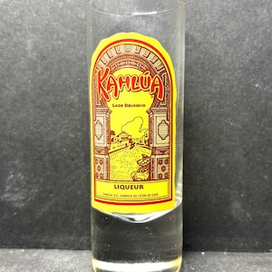 May include: A clear shot glass with a yellow and brown Kahlúa liqueur label. The label features the word "Kahlúa" in red script, with the words "Liqueur" and "Kahlúa, S.A. Fabrica de Licor de Cafe" printed on the label.