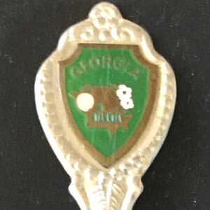 May include: Silver souvenir spoon with a green enamel shield featuring the state of Georgia and the text "Georgia" on the shield.