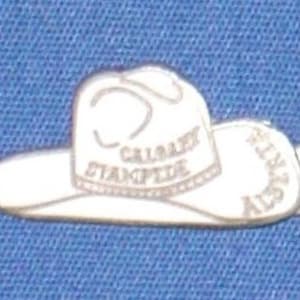 Könnte beinhalten: Weißer Emaille-Pin mit einem Cowboyhut mit dem Text "Calgary Stampede" auf dem Hutband.