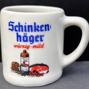 May include: A white ceramic mug featuring the blue text Schinken häger and red text würzig-mild. The graphic depicts a bottle, a glass, a loaf of bread, a piece of meat, and red berries. The mug has a sturdy handle and a smooth, cylindrical body.