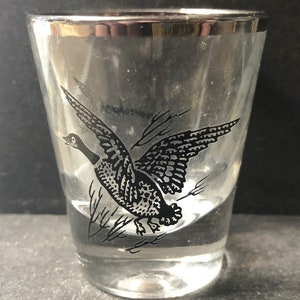 May include: A clear glass shot glass with a silver rim and a black and white image of a duck in flight.