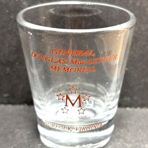 May include: Clear shot glass with red lettering that reads "GENERAL DOUGLAS MacARTHUR MEMORIAL" and "NORFOLK, VIRGINIA". The glass also features a red "M" surrounded by stars.
