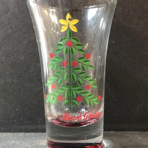 May include: A clear glass shot glass with a red rim and base. The glass features a green Christmas tree with red berries and a yellow star on top. The text "Busch Gardens" is printed in red at the bottom of the glass.
