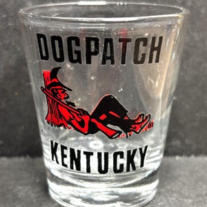 May include: Clear shot glass with the words "DOGPATCH KENTUCKY" in black block letters. A red and black graphic of a person reclining with a hat and staff is below the word "DOGPATCH".