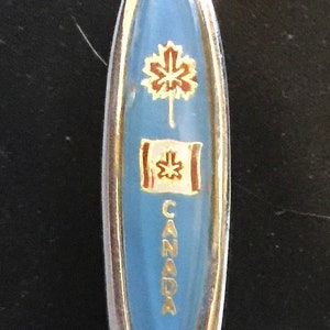Può includere: Un cucchiaio d'argento con un manico in smalto blu. Il manico presenta una foglia d'acero dorata, una bandiera canadese e la parola "CANADA" in lettere dorate.
