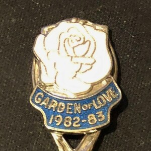 May include: A silver spoon with a blue and white enamel design. The design features a white rose and the text "Garden of Love 1982-83".