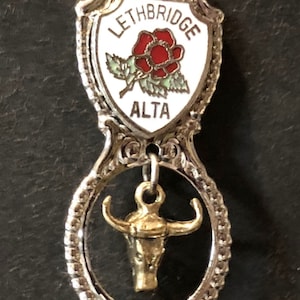 Puede incluir: Cuchara de recuerdo de plata con un dije de cabeza de toro de oro. La cuchara tiene un dije en forma de escudo con una rosa roja y el texto "LETHBRIDGE ALTA."