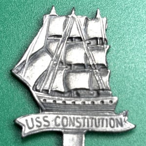 Può includere: Una spilla in metallo color argento raffigurante una nave alta con vele. La spilla presenta la scritta "USS CONSTITUTION" su uno stendardo sotto la nave. La spilla è posta su uno sfondo verde.