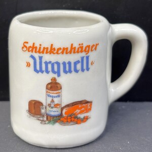 May include: A white ceramic mug featuring the orange text Schinkenhäger and blue text Urquell. Below the text is a graphic illustration of a brown bottle, a small glass, a loaf of bread, a slice of meat, and a cluster of orange berries.