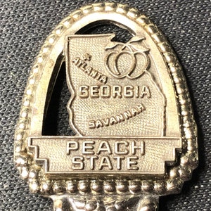 May include: A gold-colored souvenir spoon with a peach design and the words "Georgia Peach State" on it. The spoon features an outline of the state of Georgia with the cities of Atlanta and Savannah labeled.