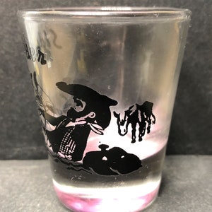 May include: A clear glass shot glass with a black silhouette of a person riding a donkey. The glass has the word "Denver" printed on the side.
