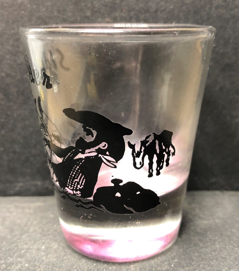 May include: A clear glass shot glass with a black silhouette of a person riding a donkey. The glass has the word "Denver" printed on the side.