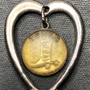 May include: Silver-toned heart-shaped pendant with a circular charm. The charm features the word "TEXAS" and an image of a cowboy boot. The pendant is likely a souvenir or a piece of jewelry.
