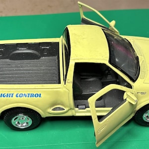 Può includere: Un camioncino giocattolo giallo con le parole "FLIGHT CONTROL" stampate sul lato. Le porte sono aperte, rivelando l'interno. Il camion ha pneumatici neri e un cassone nero.