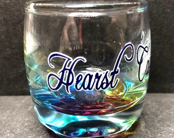 May include: A clear shot glass with a colorful, iridescent finish. The glass has the name "Hearst Castle" in a blue script font. The glass is cylindrical with a rounded base.