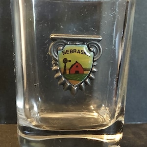 May include: A clear glass shot glass with a silver metal charm attached to the rim. The charm is shaped like a shield with the word "NEBRASKA" in blue lettering and a red barn with a windmill in a green field.