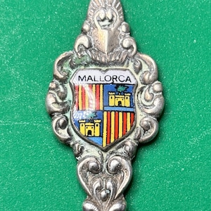 May include: A silver souvenir spoon with an ornate handle and a colorful enamel crest. The crest features the word "MALLORCA" above a shield with vertical stripes and castle illustrations. The spoon is set against a green background.