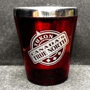 May include: A red shot glass with a silver rim. The glass features a white logo with the text "YUKON CANADA'S TRUE NORTH" and three maple leaf symbols. The shot glass is made of a transparent material.