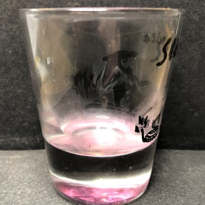 May include: A clear glass shot glass with a pink base. The glass features a black silhouette of a person wearing a hat and the word "Sea" inscribed on the side.