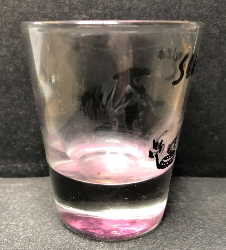 May include: A clear glass shot glass with a pink base. The glass features a black silhouette of a person wearing a hat and the word "Sea" inscribed on the side.
