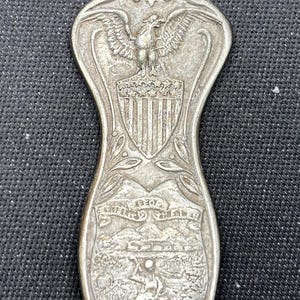 May include: Silver-toned spoon with detailed engravings. The handle features an eagle above a shield and a landscape scene with text. The spoon's design is ornate and vintage, suggesting a collectible or decorative item.