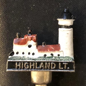 Puede incluir: Un tapón de botella de metal plateado con un diseño de faro pintado en blanco y rojo. El texto "HIGHLAND LT." está impreso en la base del tapón.