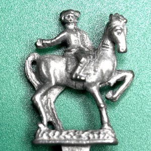 May include: A silver-coloured metal figurine of a person riding a horse. The rider wears a hat and jacket. The horse is in a trotting pose. The figurine is mounted on a base.