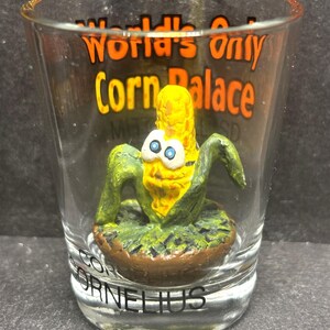 Könnte beinhalten: Ein klares Schnapsglas mit dem Text "World's Only Corn Palace" in Orange und Schwarz. Im Inneren befindet sich eine maisförmige Figur mit Gesicht, grünen Blättern und dem Namen "Cornelius" am Boden. Die Figur ist gelb, grün und braun.