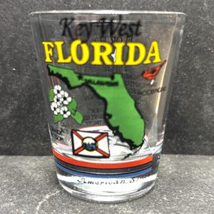 May include: A clear glass shot glass with a green outline of the state of Florida, the words "Key West Florida" in yellow lettering, and a red cardinal. The glass also features a white flower, a blue and white Florida state flag, and the words "American Souvenir" at the bottom.