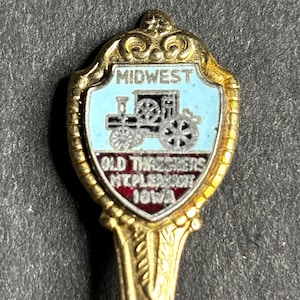 Può includere: Un cucchiaio souvenir color oro con un manico decorativo e un emblema a forma di scudo. L'emblema presenta uno sfondo azzurro con un'illustrazione di trattore nero e argento e il testo "MIDWEST" e "OLD THRESHERS MT. PLEASANT IOWA."