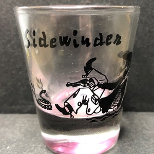 May include: A clear glass shot glass with a black and white illustration of a cowboy riding a snake with the word "Sidewinder" printed on the glass.