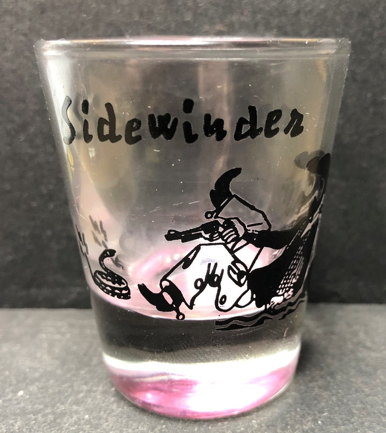 May include: A clear glass shot glass with a black and white illustration of a cowboy riding a snake with the word "Sidewinder" printed on the glass.