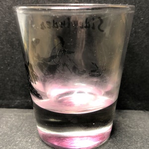 May include: A clear glass shot glass with a pink tint at the bottom. The glass has a black silhouette of a bird and a snake etched on the side. The text "Sidewalk" is etched on the top of the glass.