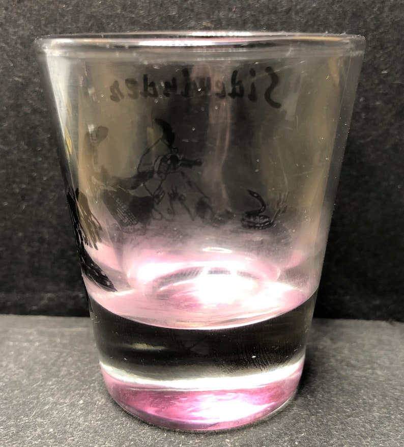 May include: A clear glass shot glass with a pink tint at the bottom. The glass has a black silhouette of a bird and a snake etched on the side. The text "Sidewalk" is etched on the top of the glass.